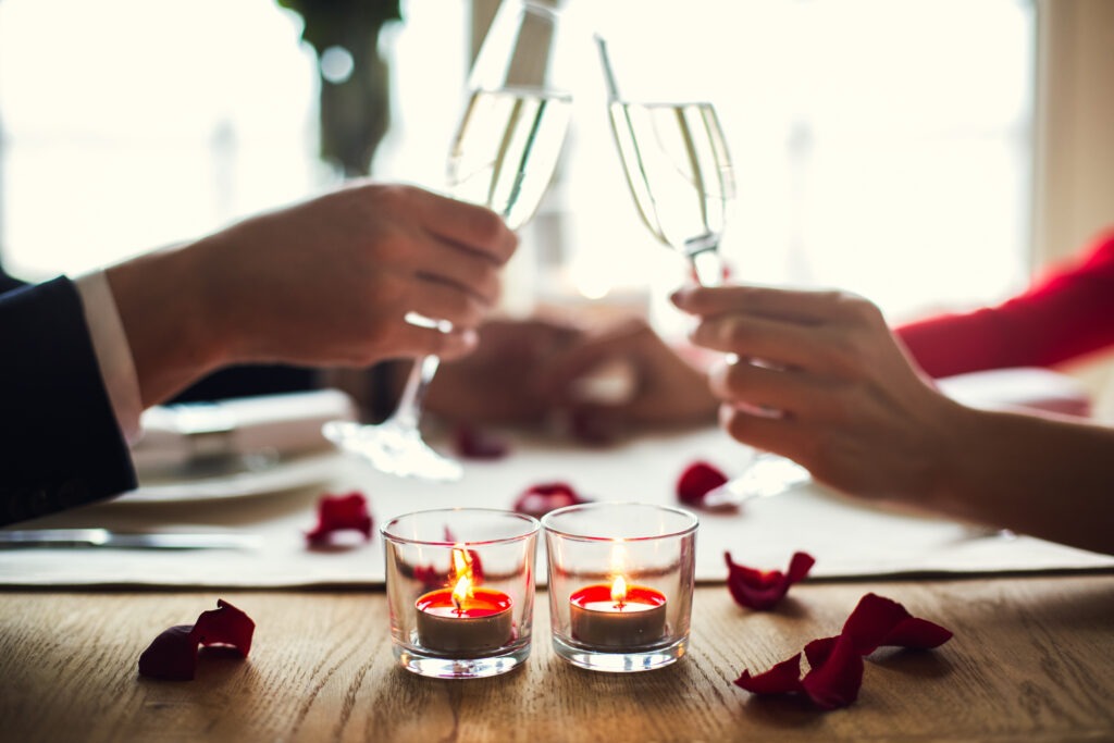 Young man and woman having romantic dinner in the restaurant holding champagne glasses toast close-up rose petals on the table